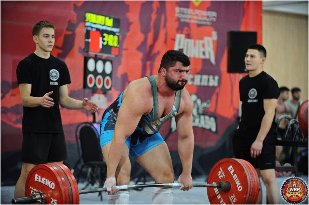 Mann hebt eine Hantelstange bei einem Gewichtheben-Wettkampf bei den 2018 World Weightlifting Championships, umgeben von Zuschauern und Offiziellen in einer Sporthalle.