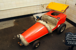 Ein rotes Pedalauto auf einem Parkplatz neben einem Schild geparkt, mit Rohren an der Wand im Hintergrund.