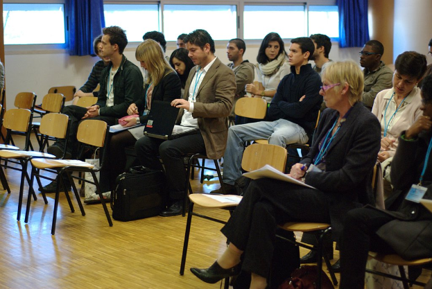 Mehrere Personen sitzen auf Stühlen in einem Raum, einige halten Gegenstände wie Laptops und Stifte, mit leeren Stühlen, Fenstern und Vorhängen.