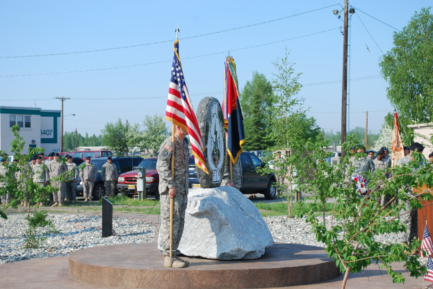 Zwei Militärangehörige halten Fahnen auf beiden Seiten eines Gedenksteins, mit Pflanzen, Autos, anderen Militärangehörigen, Strommasten und Gebäuden im Hintergrund.