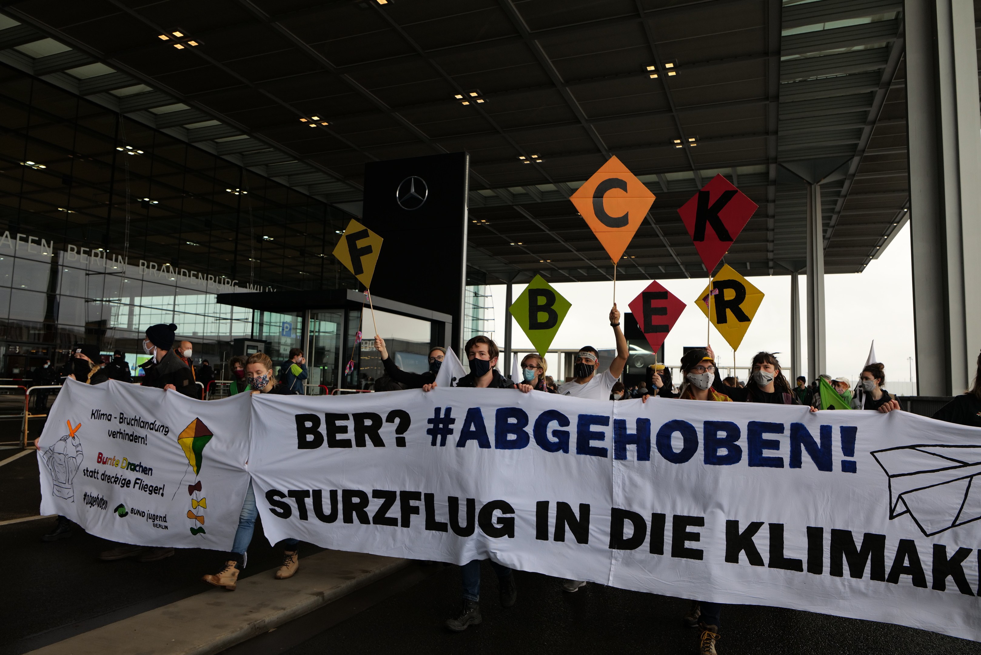 People wearing masks hold a banner reading "Ber? Abgehoben Sturzflug in die Klimakt" outside a building with glass walls and illuminated pillars.