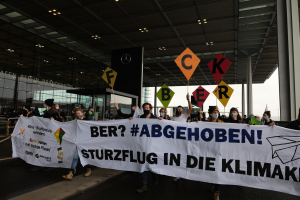 People wearing masks hold a banner reading "Ber? Abgehoben Sturzflug in die Klimakt" outside a building with glass walls and illuminated pillars.