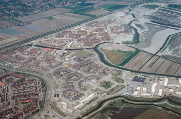 Luftaufnahme einer Stadt mit Gebäuden, Straßen, Feldern, Bäumen und einem zentralen Reservoir, umgeben von städtischen und ländlichen Gebieten.