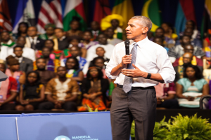Präsident Obama spricht auf einer Wahlveranstaltung, hält ein Mikrofon, mit einer Menge von Menschen, einige sitzen und einige stehen, eine Pflanze im Vordergrund und Flaggen und eine Tafel im Hintergrund.