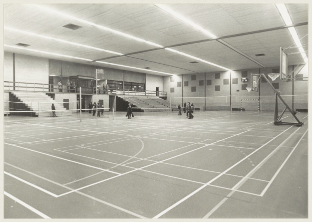Schwarzes und weißes Foto einer Turnhalle mit Menschen, die Badminton auf einem Court spielen, Treppe mit Geländer, Deckenlampen und Basketballkorb auf der rechten Seite.