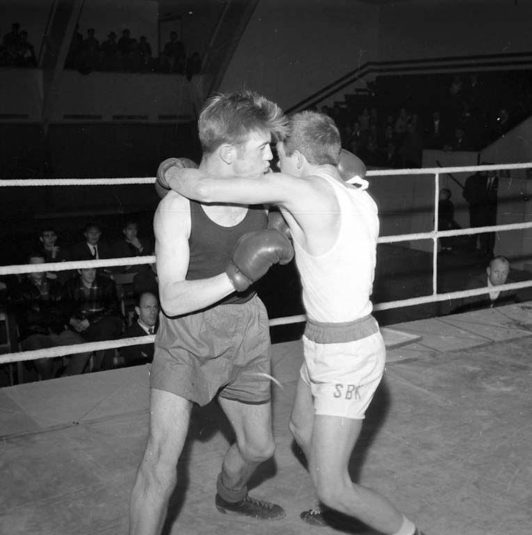 Zwei Boxer in einem Boxring umgeben von sitzenden und stehenden Zuschauern in Schwarz-Weiß.