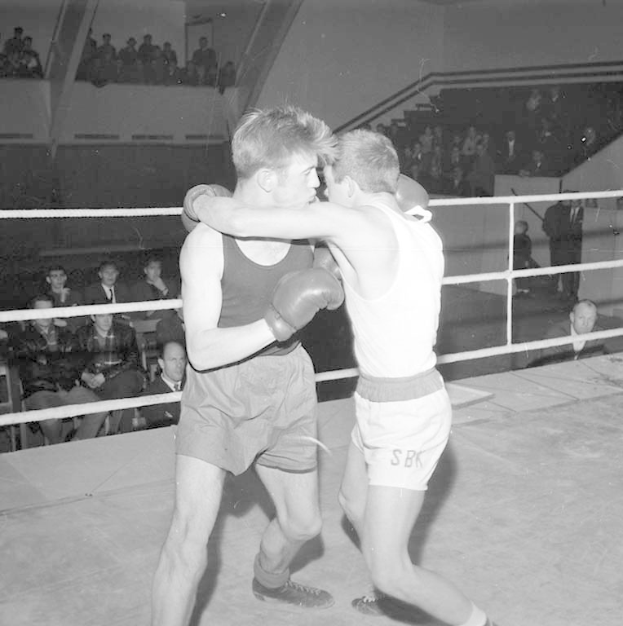 Zwei Boxer in einem Boxring umgeben von sitzenden und stehenden Zuschauern in Schwarz-Weiß.