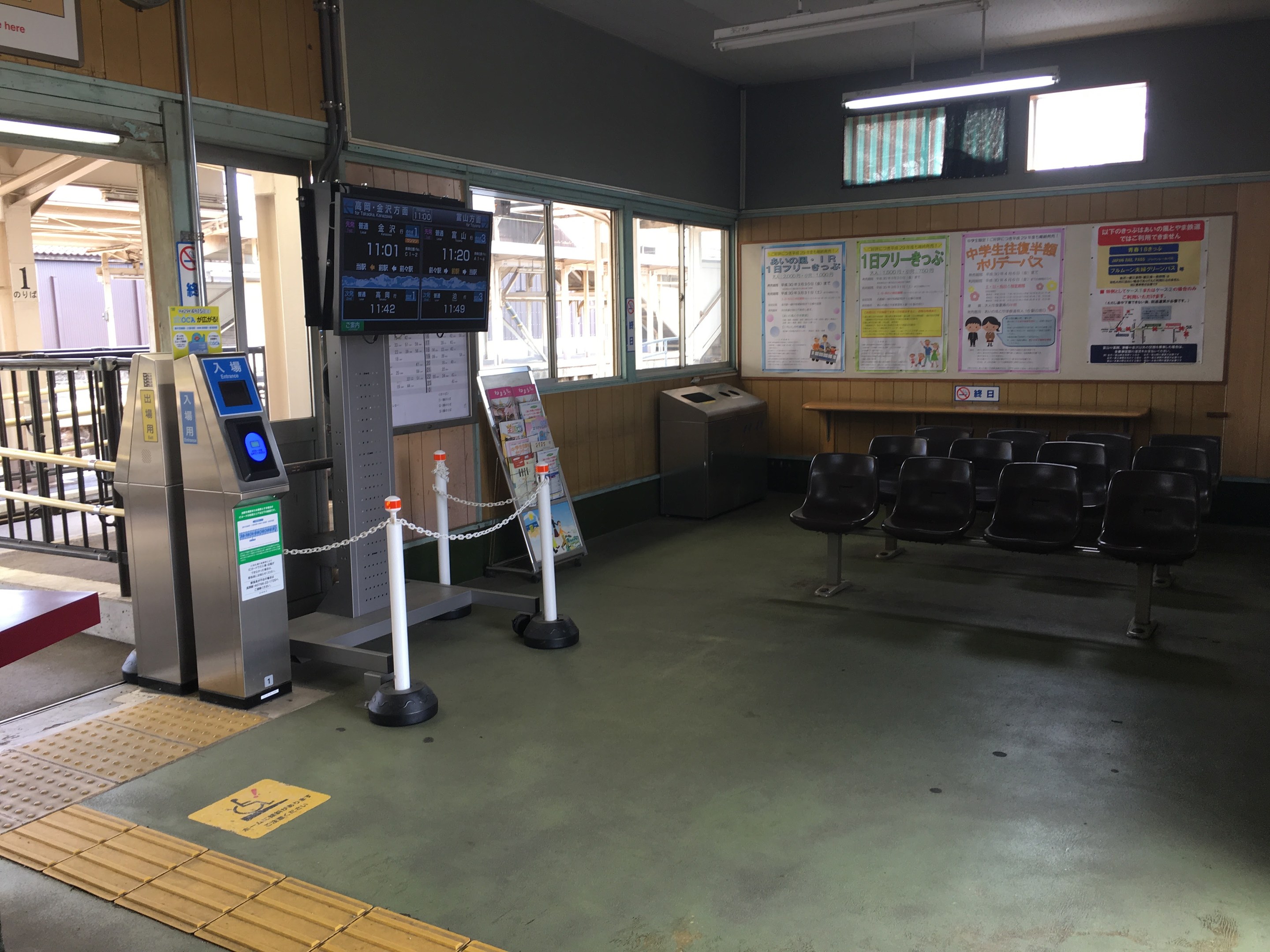 Ein belebter Bahnhofseingang mit Sitzgelegenheiten, einer Informations counter, Ticketautomaten, Deckenleuchten, Geländern und informativen Tafeln und Plakaten an den Wänden.