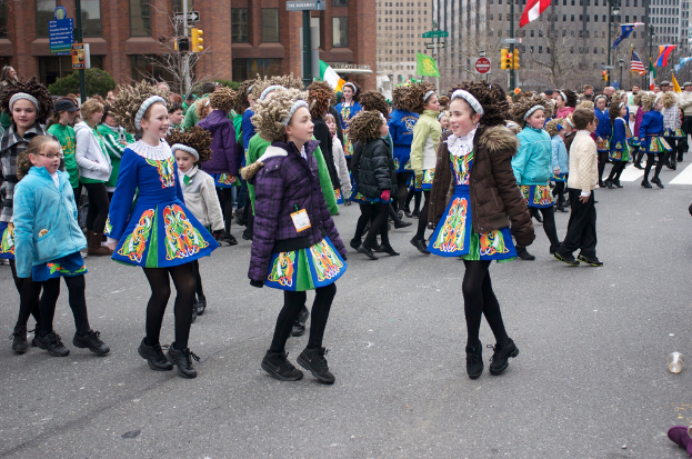 Eine Gruppe von Mädchen in Kleidern mit Jacken und Pullover, die auf einer Straße tanzen, mit einem roten Gebäude und einem gelben Signalmast dahinter und Gebäuden mit Flaggen auf Strommasten im Hintergrund.