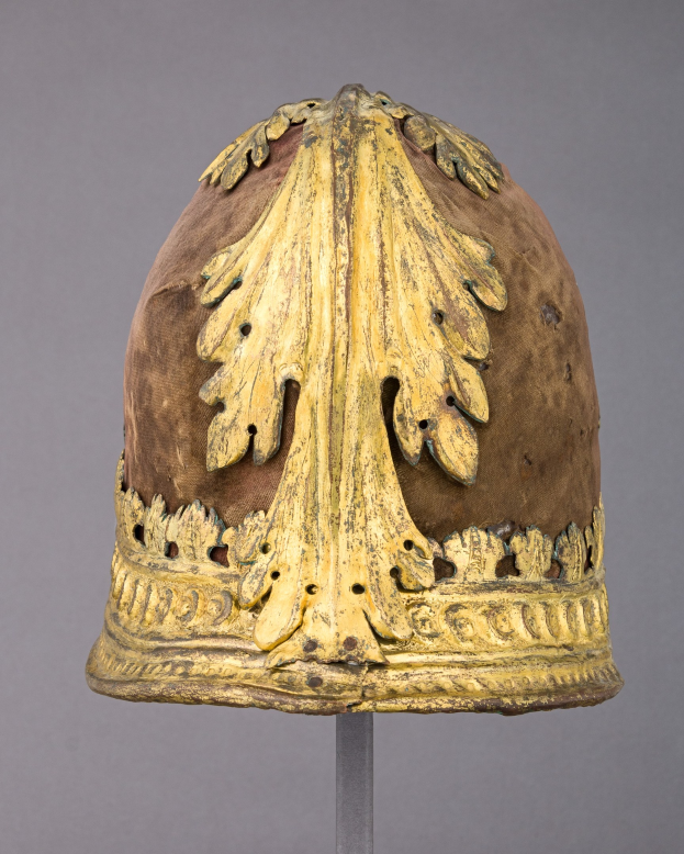 Goldener Helm mit Krone und detaillierten Verzierungen auf einem Sockel vor einem grauen Hintergrund.
