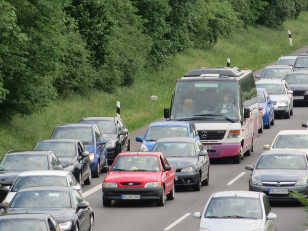 Ein Stau auf der Autobahn mit zahlreichen Autos und einem Lieferwagen, Menschen in den Fahrzeugen sichtbar, Bäume und Gras im Hintergrund.