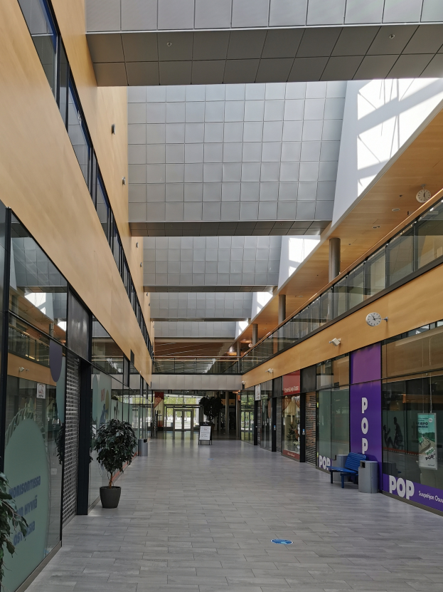 Das Innere eines großen Gebäudes mit vielen Fenstern, Hauspflanzen, einer Bank, einem Müllkorb, Tafeln mit gedrucktem Text, Glastüren, Lichtern und anderen Gegenständen.