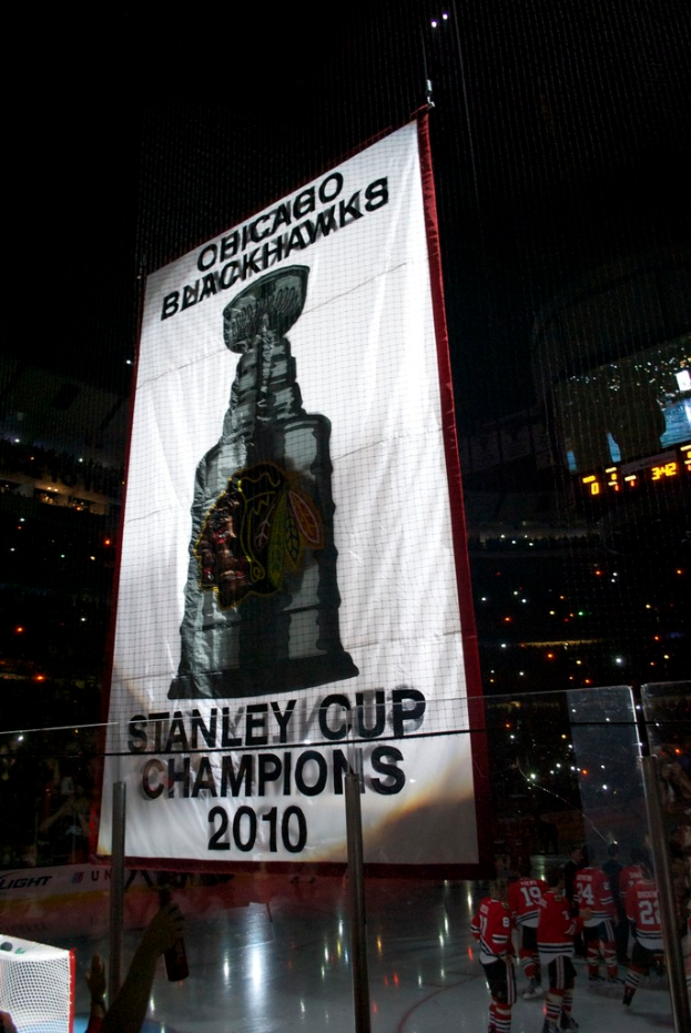 Eine nächtliche Stadion-Szene mit einem großen Banner in der Mitte, Zuschauern im Hintergrund, einem Glas auf dem Boden, einigen Spielern auf dem Feld und beleuchteten Rängen.