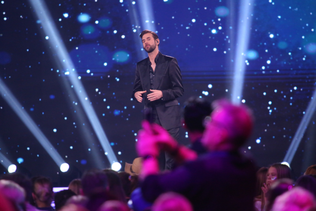 A man in a black suit stands on a brightly lit stage, holding a trophy, identified as the winner of *Dancing with the Stars* in front of a crowd.