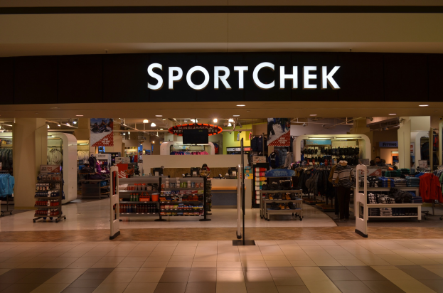 Außen- und Innenansicht eines Sportchek-Einzelhandelsgeschäfts in einem Einkaufszentrum, mit Kleiderstangen, Deckenleuchten, Säulen und einem Fernseher.