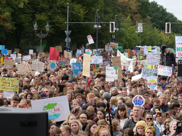 Eine große Menschenmenge mit Schildern steht vor einem Gebäude unter einem klaren blauen Himmel, mit Laternen, Verkehrsampeln und Bäumen im Hintergrund; einige tragen Mützen, Schutzbrillen oder halten Kameras, und ein schwarzes Objekt ist in der unteren linken Ecke sichtbar.