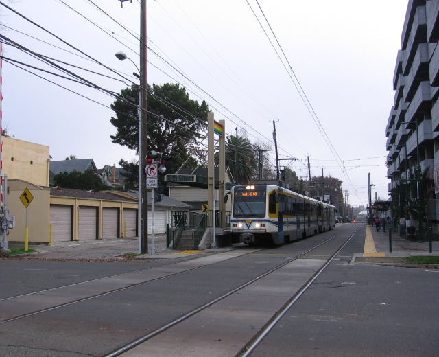 Eine Straßenbahn fährt auf einer Straße mit hohen Gebäuden auf beiden Seiten, Strommasten mit Oberleitungen, Fußgängern auf dem Gehweg, Bäumen im Hintergrund und einem klaren blauen Himmel.