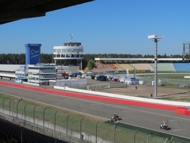 Eine Gruppe von Menschen auf Motorrädern auf einer Rennstrecke umgeben von einem Metallzaun und anderen Strecken-Elementen unter einem bewölkten Himmel.