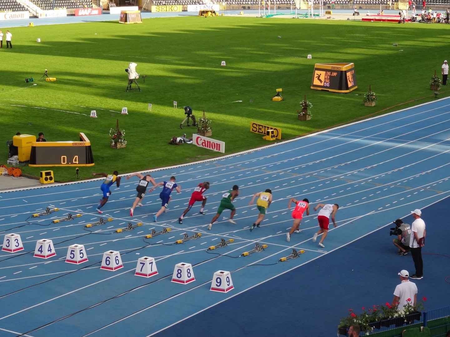 Group of people running on an outdoor track in a stadium with a fence, flowered plants, text boards, camera equipment, chairs, and poles against a clear blue sky.