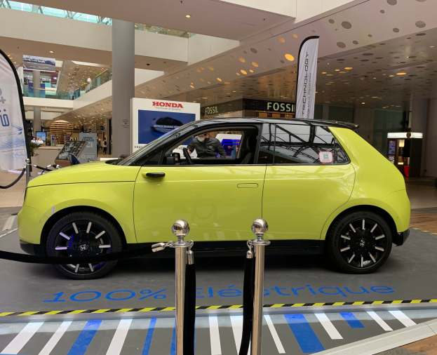 Gelber wasserstoffbetriebenes elektrisches Auto vor einem Gebäude mit Absperrpfosten, Bannern, Menschen, Schildern, einer Topfpflanze, Deckenleuchten und Ventilatoren.