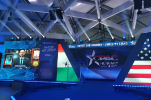 Eine Bühne für ein politisches Ereignis mit einer amerikanischen Flagge, einer Person an einem Rednerpult mit einem Mikrofon, Tafeln mit Text und Bildern, einem Bildschirm mit einer Person, Deckenlampen und Stangen sowie CPAC Hungary-Wasserzeichen.