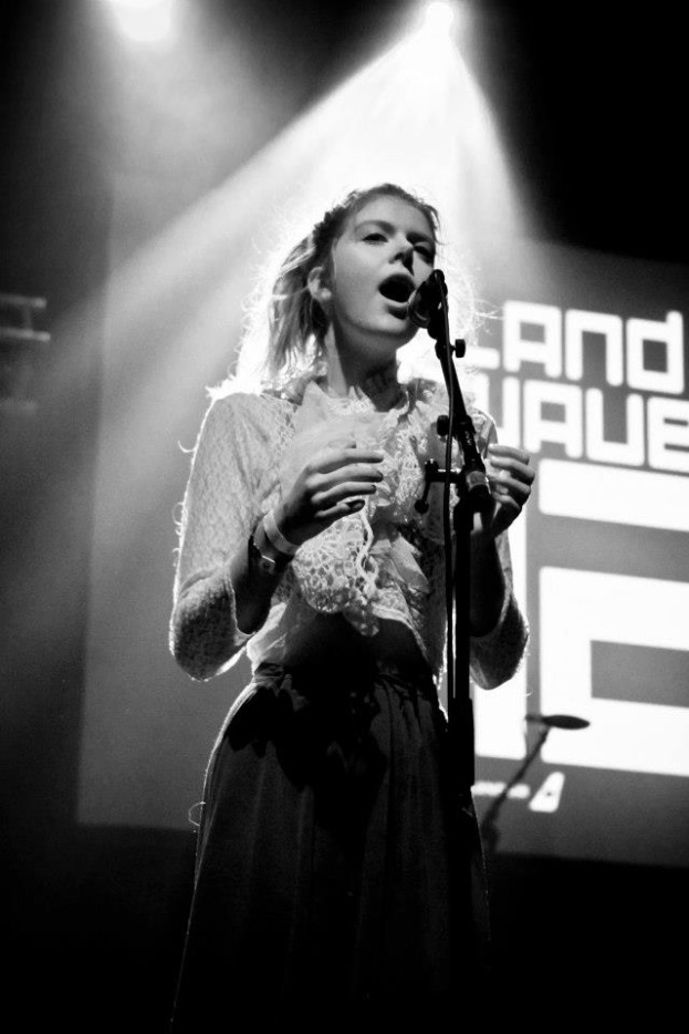 Eine Frau steht auf einer Bühne, singt in ein Mikrofon mit Mikrofonständer vor sich, beleuchtet von hellen Lichtern, mit einem Bildschirm dahinter, in einem Schwarz-Weiß-Bild.