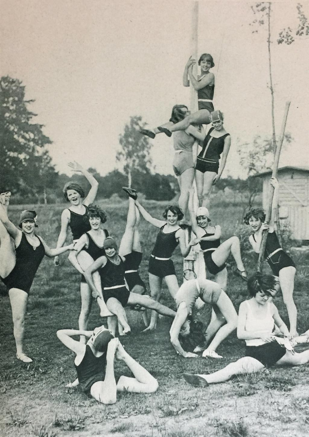 Eine Gruppe von Frauen in Badeanzügen posiert für ein Foto auf einer grünen Wiese umgeben von Bäumen und einem Haus im Hintergrund, schwarz-weiß, 1920er Jahre.