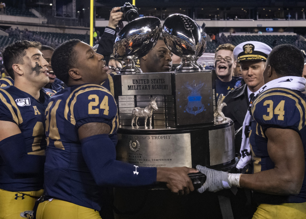 A group of men celebrating a victory, with one holding a trophy and another taking a photo, in a brightly lit stadium with a cheering crowd and poles in the background.