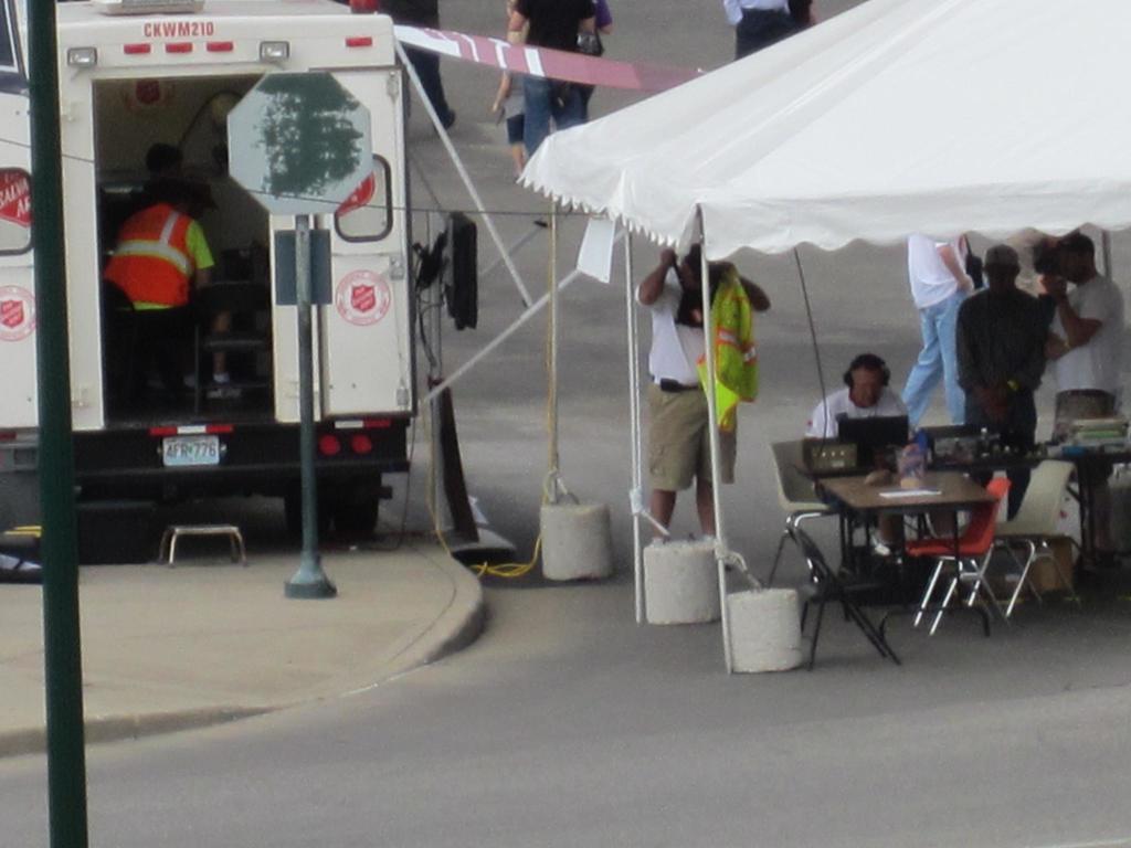 Ein Rettungswagen steht auf der linken Seite einer Stra├če, w├Ąhrend auf der rechten Seite ein Zelt viele Menschen sch├╝tzt, mit einem Tisch und einem Stuhl in der N├Ąhe.