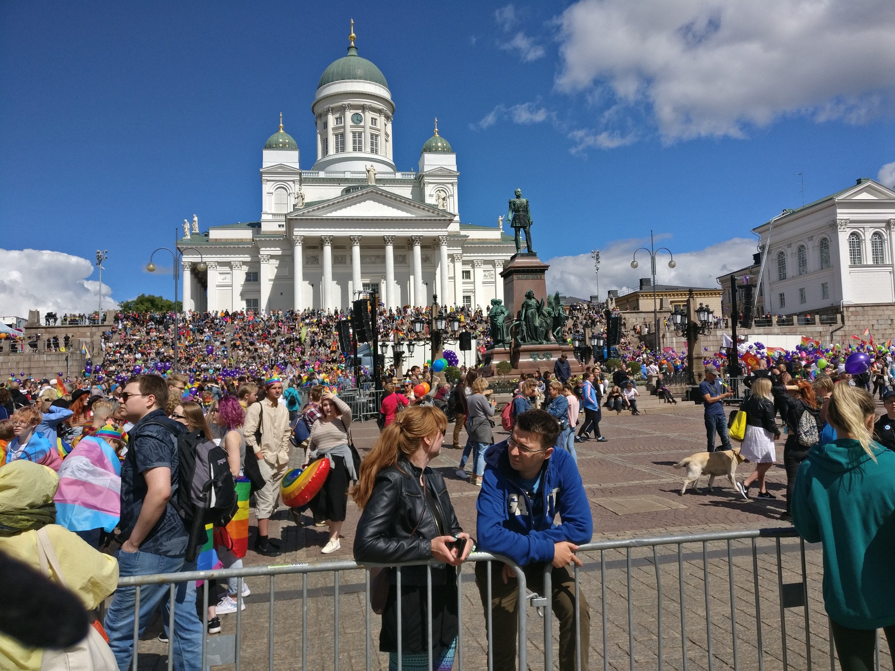 Large crowd gathered outside a building with pillars and arches during a Helsinki pride parade, some holding balloons and wearing bags.