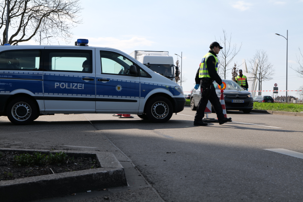 Ein Polizeifahrzeug steht auf der Seite einer Straße neben einem Verkehrskegel, mit Fahrzeugen, Kegeln, Gras, Bäumen, Laternenmästen, Schildtafeln, Gebäuden und einem bewölktem Himmel im Hintergrund.