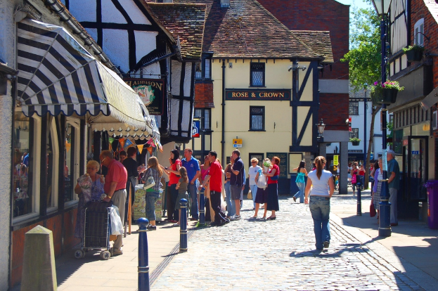Eine Straße mit Menschen auf der linken Seite neben einem Handwagen und einem Mann und einer Frau auf der rechten Seite neben einem lila Müllcontainer, mit Gebäuden, Bäumen und Blumentöpfen im Hintergrund.