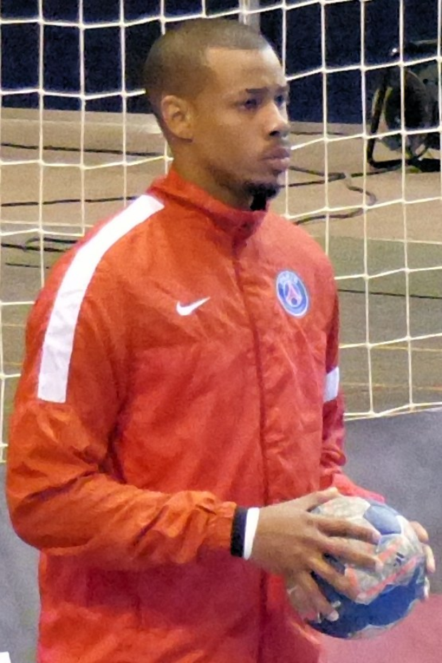 Ein Mann in einem blauen T-Shirt und Shorts steht vor einem Tor, hält einen Fußball und hat einen entschlossenen Gesichtsausdruck.