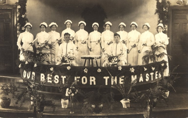 Eine Gruppe von Frauen in weißen Kleidern und Hauben steht auf einer Bühne mit einem Banner, auf dem 'Unser Bestes für den Meister' steht, umgeben von Blumentöpfen, mit Türen und einer Wand im Hintergrund.