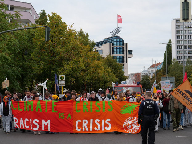 Gruppe von Menschen marschiert mit einem "Klima-Krise ist eine Krise"-Schild die Straße entlang, mit Bäumen, Laternenmasten und geparktem Fahrzeug an der Straße, Gebäude im Hintergrund unter einem klaren blauen Himmel.