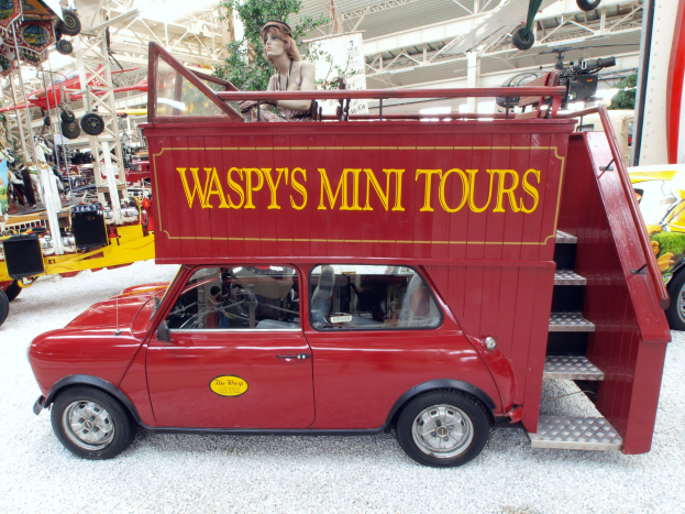 Ein Kleiner Roter Wagen Mit Der Aufschrift "Waspy's Mini Tours" Steht Zwischen Anderen Fahrzeugen Und Gegenständen In Einem Innenraum.
