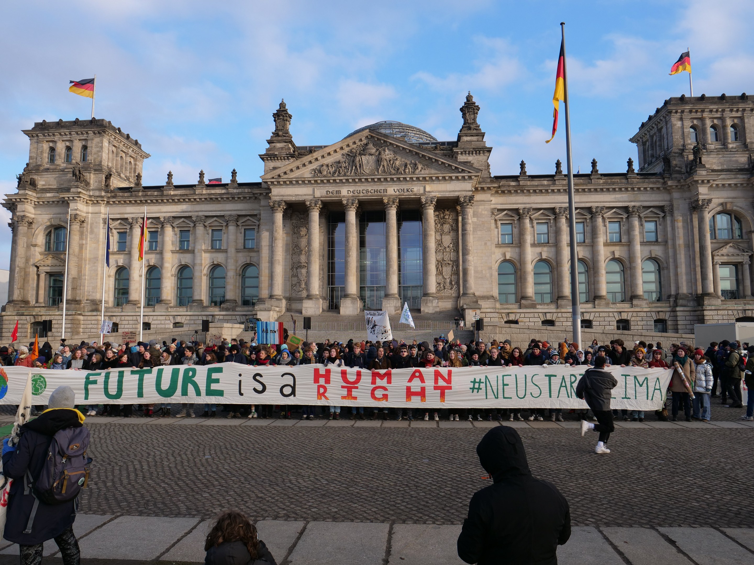 A group of people holding a banner with the text "Future is a Human Neustar ima" in front of the Reichstag building in Berlin, Germany, with the building's architectural details visible and flags in the background under a cloudy sky.