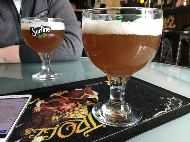 Zwei Gläser goldenes Bier auf einem Tisch mit einem Handy in der Nähe, umgeben von Menschen, mit gerahmten Kunstwerken an der Wand im Hintergrund.