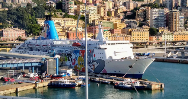 Großes Kreuzfahrtschiff liegt in der Nähe einer Stadt mit einer Brücke links, Fahrzeugen auf der Straße und Gebäuden und Bäumen im Hintergrund.