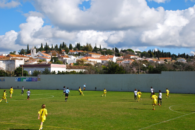 Eine Gruppe von Menschen spielt ein Spiel auf einem Feld mit Plakaten an einer Wand, Bäumen, Pfählen, Häusern und Wolken im Hintergrund.
