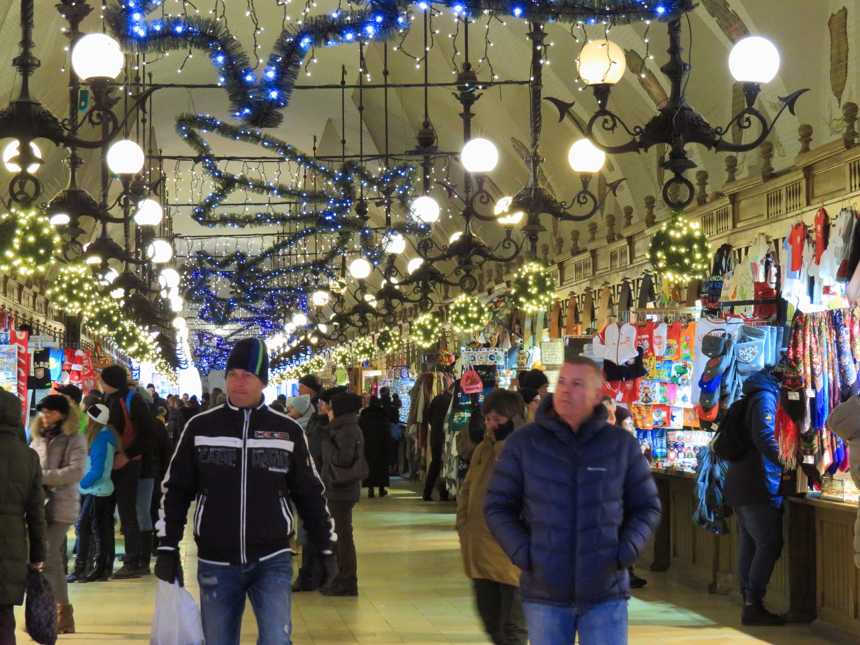 A bustling Christmas market with people walking, some wearing caps and carrying bags, under hanging lights and decorations, with clothing and goods displayed on the right and a wall in the background.