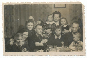 Ein Schwarz-Weiß-Foto einer lachenden Familie, die sich um einen Tisch mit Gläsern, Flaschen, Tellern und anderen Gegenständen versammelt hat, mit einem Vorhang und einem Rahmen an der Wand im Hintergrund.
