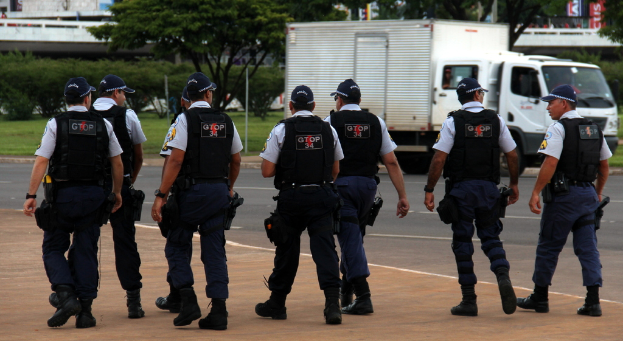 Eine Gruppe von Beamten in Uniform, einschließlich Mützen, Hemden, Schutzwesten, Uhren, Hosen und Schuhen, die auf einer Straße mit einem geparkten Lastwagen auf der rechten Seite neben einem Zaun, Gebäuden, Bäumen, Pflanzen, Polen und Bannern im Hintergrund und einer Brücke oben rechts gehen.