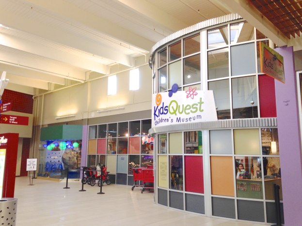 Innenseite eines Einkaufszentrums mit einem "Kids Quest Children's Museum"-Schild, Trolleys, buntem Band an Stangen, Informationsschildern, Deckenleuchten und verstreuten Gegenständen.
