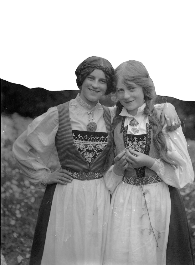 Zwei junge Frauen in traditioneller bayrischer Tracht, mit weißen und schwarzen Kleidern mit aufwändiger Stickerei und Perlen, die vor der Kamera stehen und lächeln, vor einem Hintergrund aus üppigen grünen Pflanzen und einem hellen weißen Himmel.