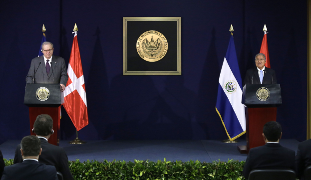 Zwei Männer stehen an Rednerpulten und sprechen zu einer sitzenden Menge, wobei der Mann links als Chilens Präsident Juan Manuel Santos identifiziert wird, flankiert von drei Flaggen und einer blauen Wand mit einem gerahmten Objekt im Hintergrund.