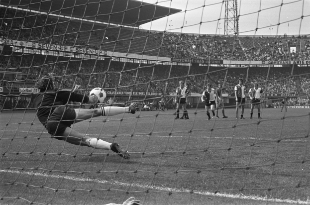 Ein Schwarz-Weiß-Foto eines Fussballspielers in Sportkleidung bei einem gehaltenen Ball vor einem Publikum in einem Stadion mit sichtbaren Rängen, Texttafeln, einem Turm und dem Himmel.