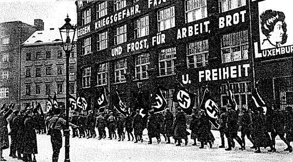 A protest march in Berlin with people holding signs and riding bicycles, passing a building with arched windows, pillars, and sculptures, set against trees and a light pole.