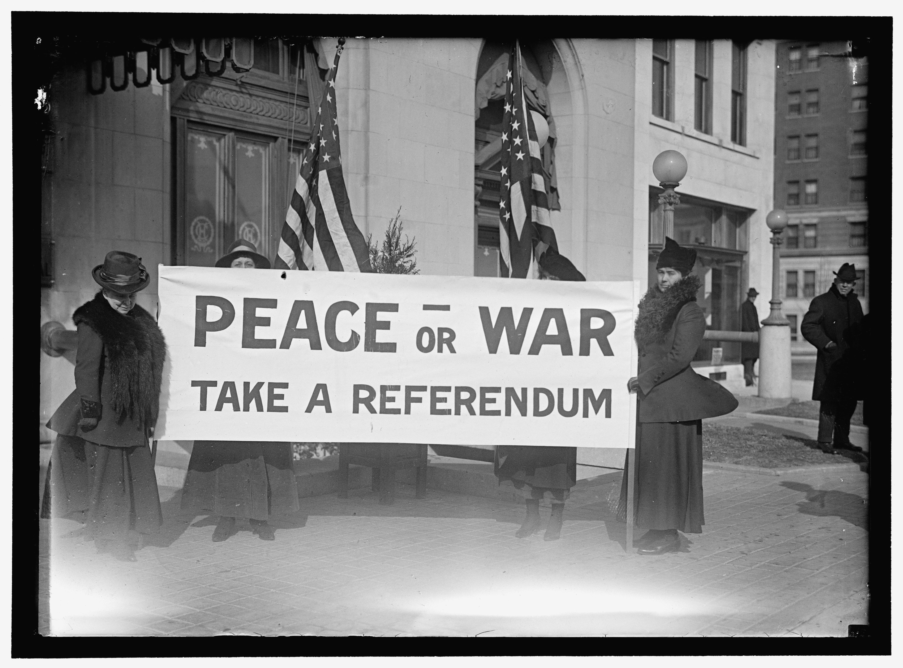 Gruppe von Frauen mit einem Banner, auf dem "Frieden oder Krieg, Abstimmung" steht, vor einem Gebäude, einer Pflanze, einem Straßenpfahl und einem bewölkten Himmel.
