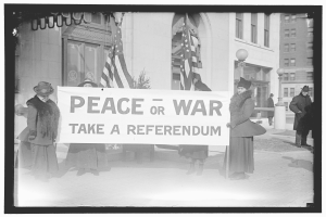 Gruppe von Frauen mit einem Banner, auf dem "Frieden oder Krieg, Abstimmung" steht, vor einem Gebäude, einer Pflanze, einem Straßenpfahl und einem bewölkten Himmel.